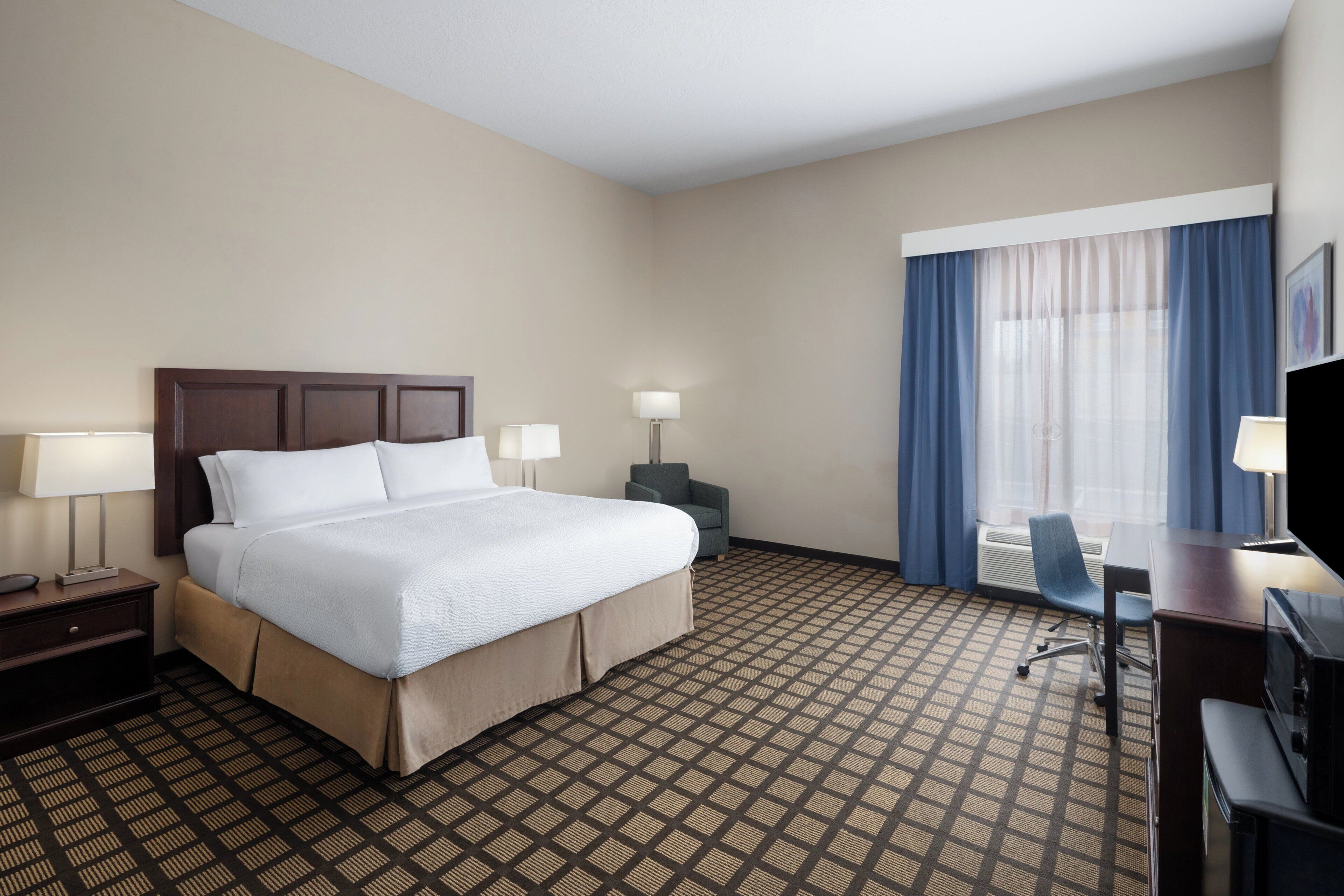 A hotel room with a bed, two bedside tables with lamps, a desk with a chair, and a large window with curtains.