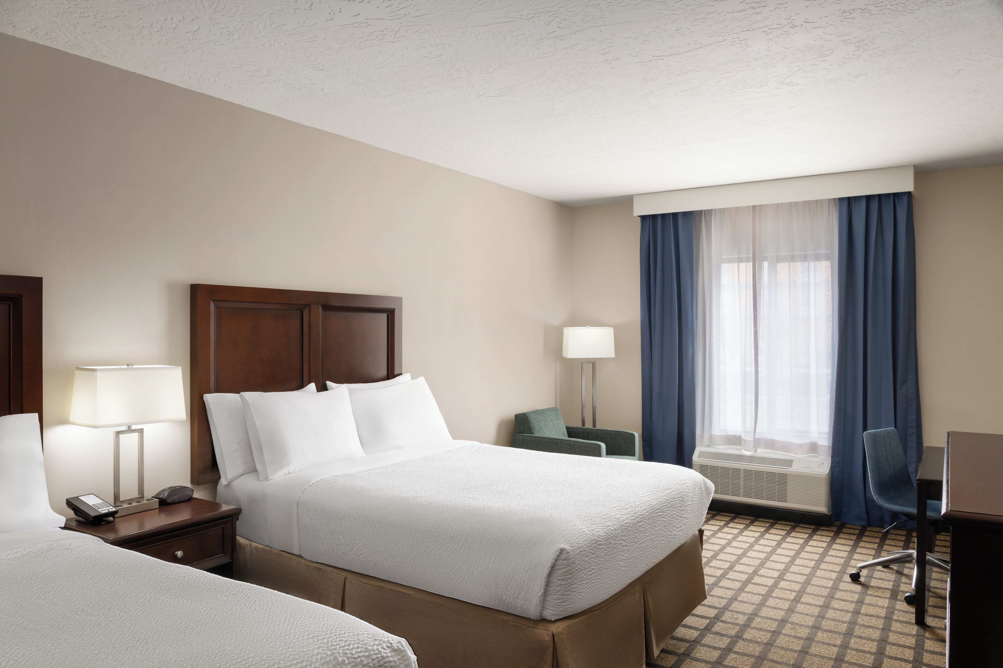 A hotel room with two beds, a desk, a chair, a lamp, and a window with curtains.