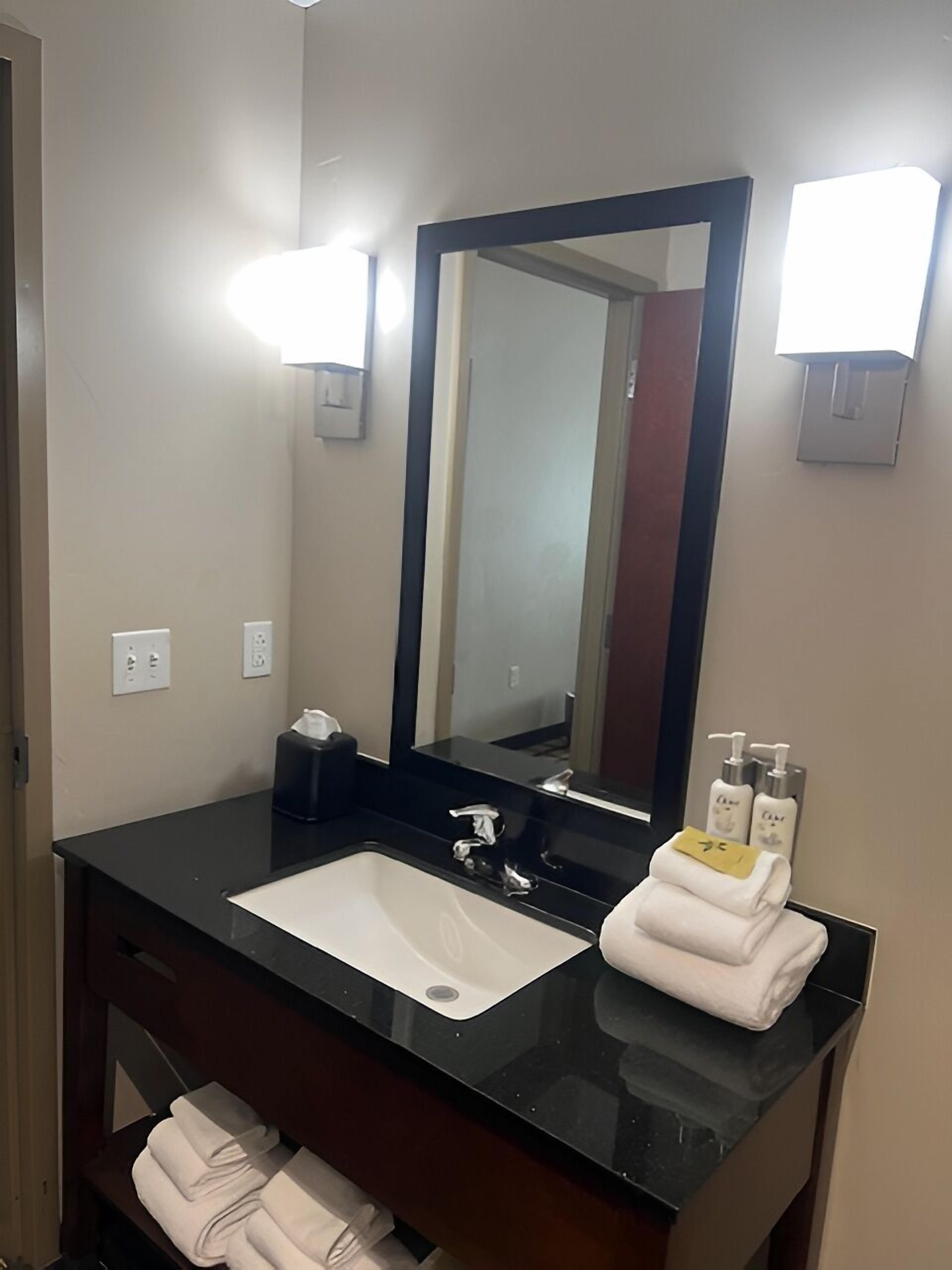 A hotel bathroom with a black countertop, a white sink, a large mirror, and two wall-mounted light fixtures.