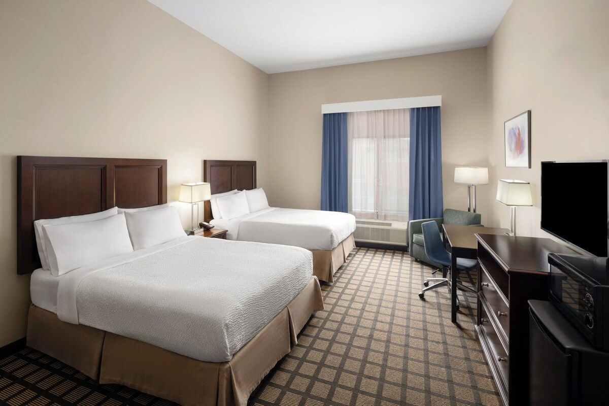 A hotel room with two beds, a desk, a TV, and a window with curtains.