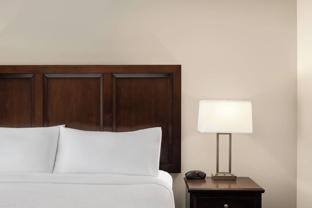 Een bed met witte lakens, een houten hoofdbord, een nachtkastje met een lamp en een klein elektronisch apparaat.