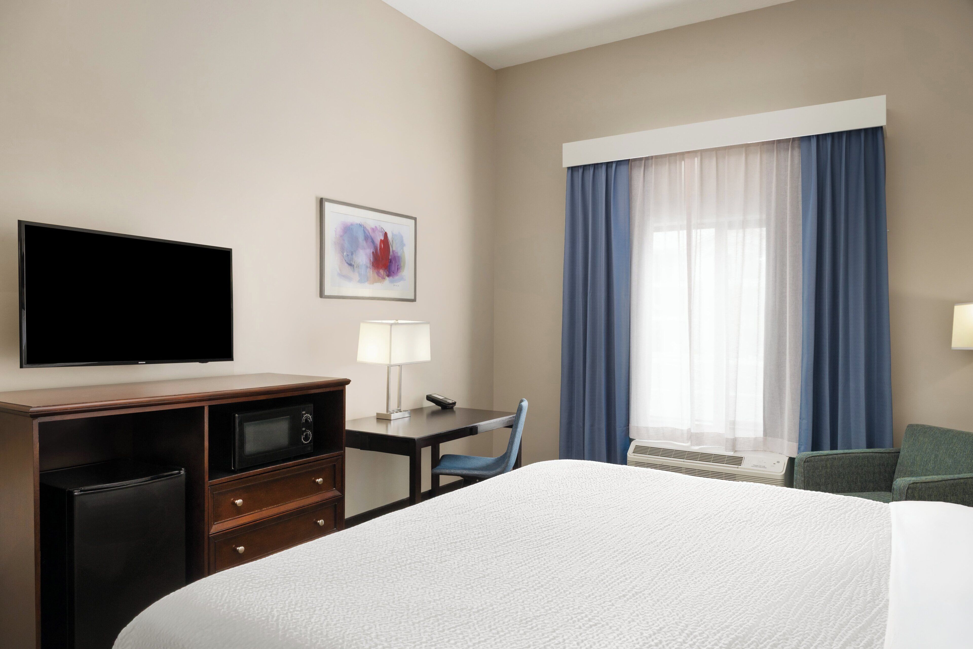 Quarto de hotel com cama, escrivaninha com luminária, televisão, micro-ondas e uma cadeira.