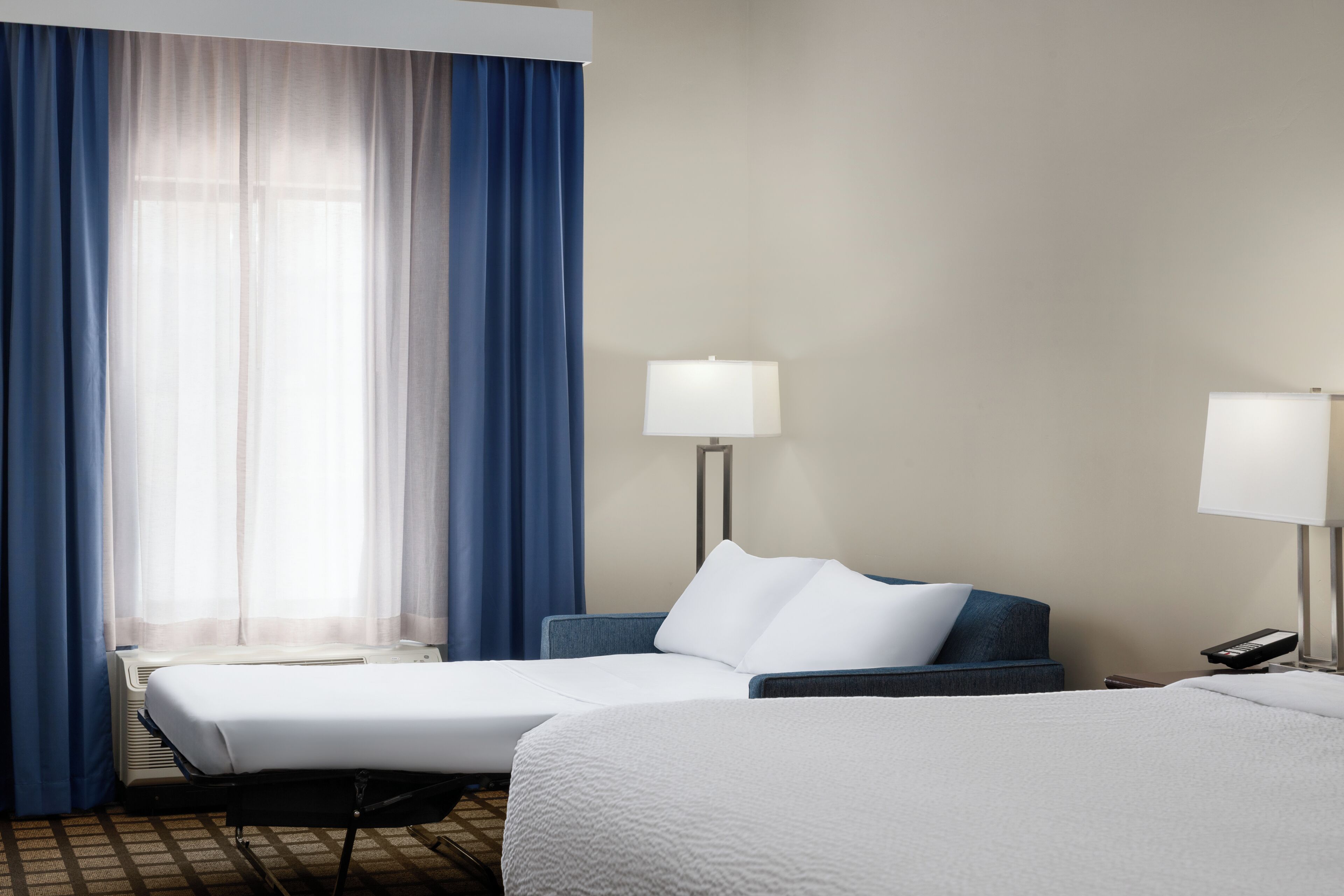 Habitación de hotel con dos camas, un escritorio, una silla, una lámpara y una ventana con cortinas.