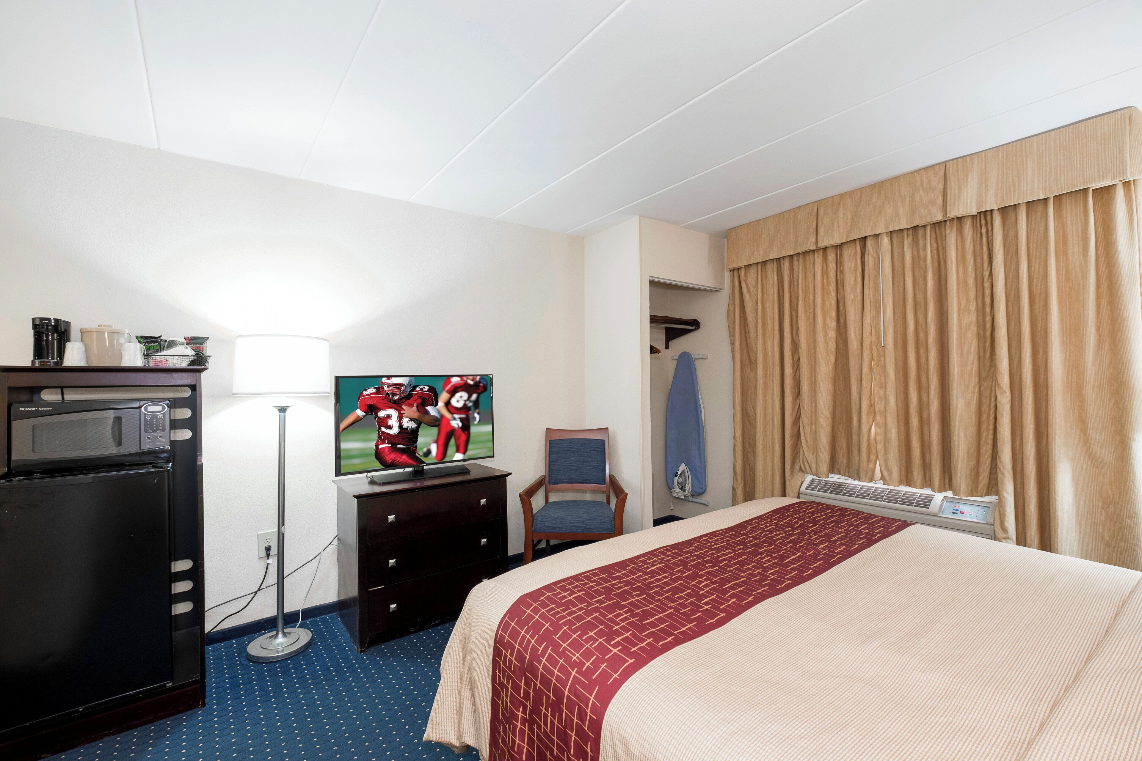 A hotel room with a bed, a desk with a computer, a microwave, a coffee maker, and a TV displaying a sports game.
