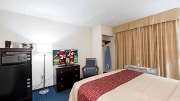 A hotel room with a bed, a desk with a computer, a microwave, a coffee maker, and a TV displaying a sports game.