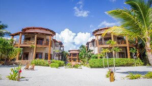 Hotel frente a la playa con edificios de dos pisos, una playa arenosa en primer plano y palmeras.
