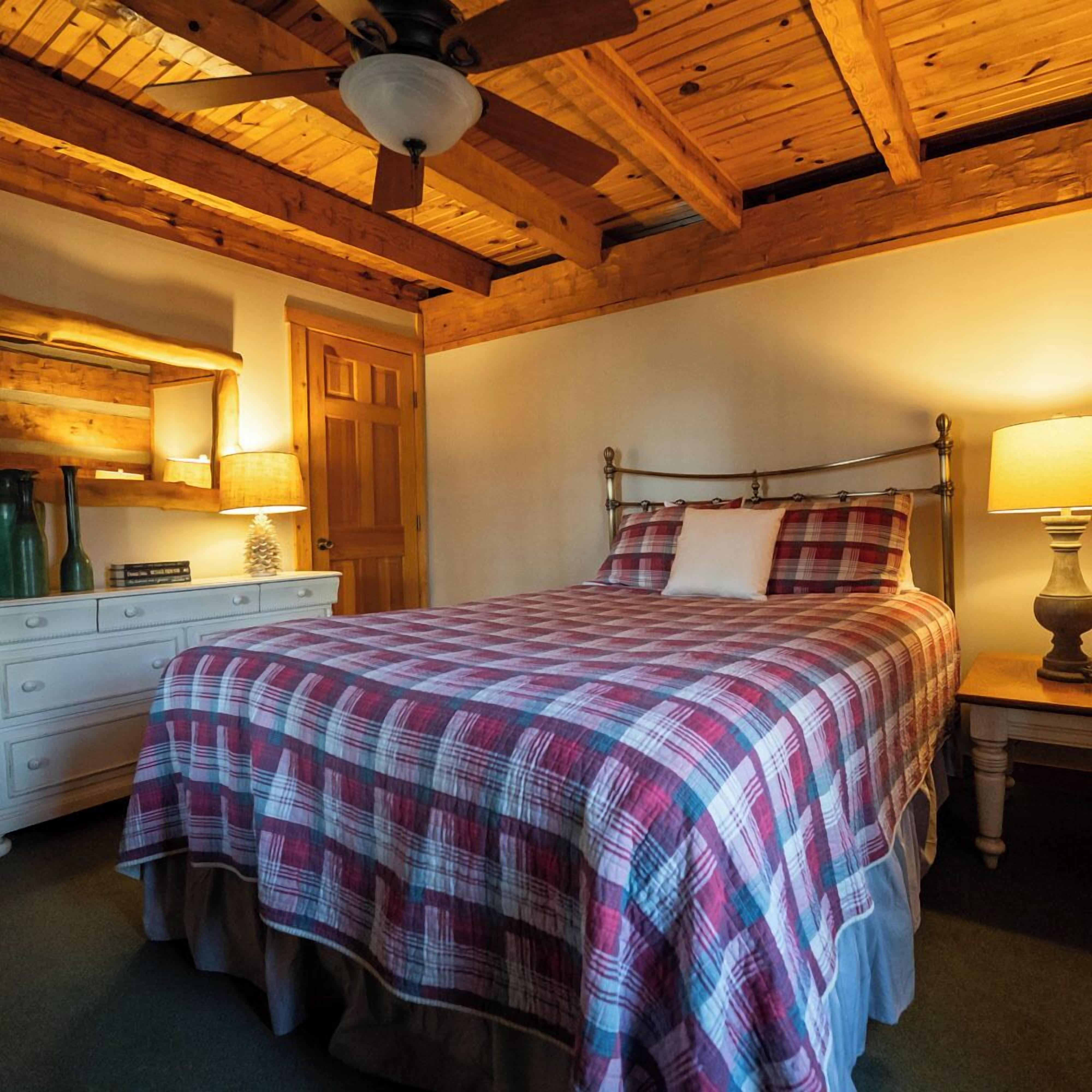 A bedroom with a wooden ceiling, a bed with a plaid bedspread, a nightstand with a lamp, and a dresser with a mirror.