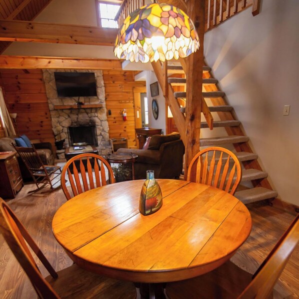 A wooden dining table with chairs, a stained glass lamp, and a stone fireplace in a cabin-style room.
