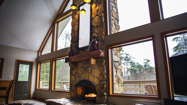 A stone fireplace with a wooden mantel, large windows, and a view of trees. - Fireside Lodge - Hosted by The Chalets (Logan)