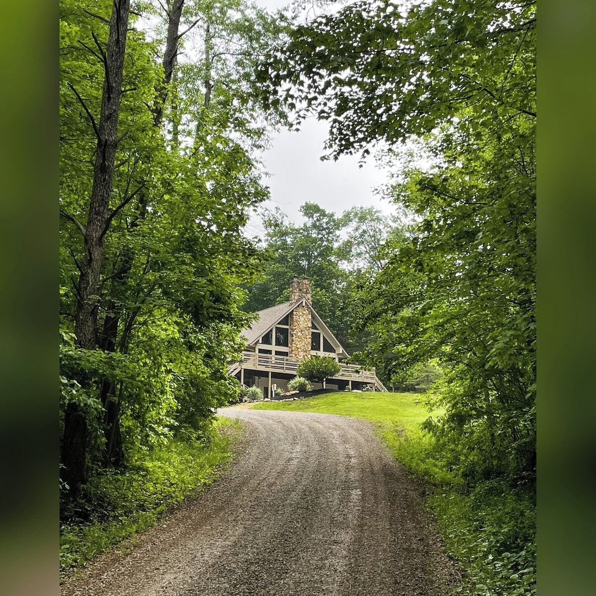 A rustic lodge with a stone chimney, surrounded by dense green foliage and a gravel path leading to the entrance.