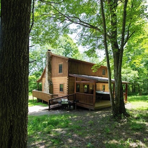 Escape to a secluded cabin surrounded by a lush forest, featuring a wooden deck and stone chimney.