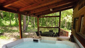 A hot tub with a wooden deck and chairs, surrounded by trees.