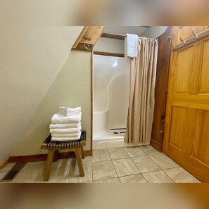 A bathroom with a shower, a stack of white towels on a wooden stool, and a wooden door.