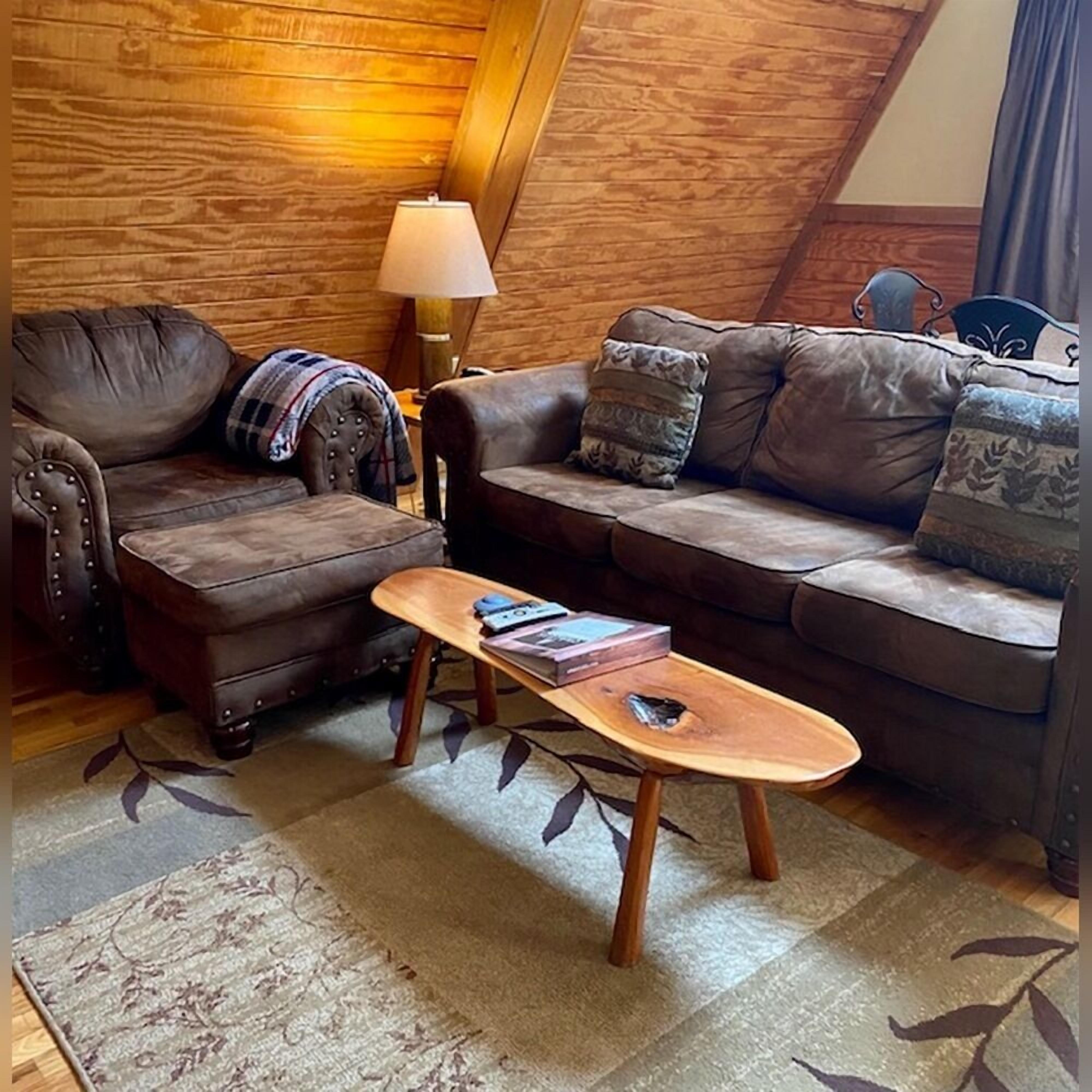 A cozy loft-style living room with a brown leather sofa, a matching armchair, and a wooden coffee table. A lamp and a patterned rug are also visible.