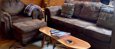A cozy loft-style living room with a brown leather sofa, a matching armchair, and a wooden coffee table. A lamp and a patterned rug are also visible.