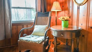 Une pièce aux murs lambrissés, avec un fauteuil à bascule, une petite table surmontée d’une lampe et un tableau encadré accroché au mur.