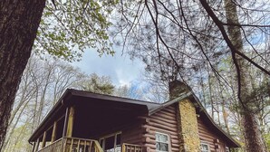 Une cabane rustique en rondins de bois, dotée d’un porche et d’une cheminée en pierre, entourée d’arbres.