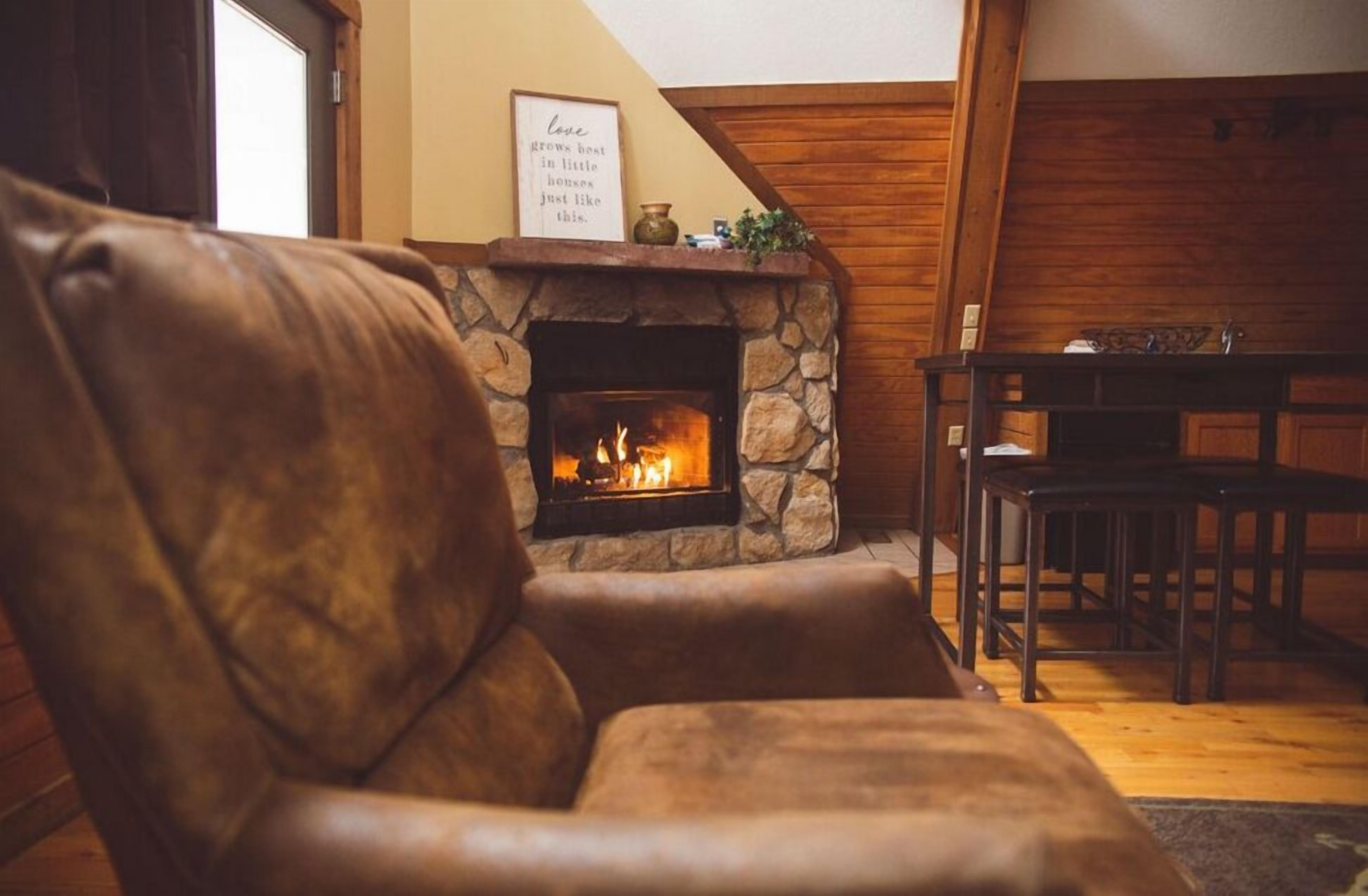 A cozy living room with a stone fireplace, leather armchair, wooden dining table, and a framed quote on the wall.