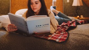 Eine Person liegt in einem Zimmer mit schräg stehender Decke auf einem Bett und liest ein Buch mit dem Titel „Van Life“.