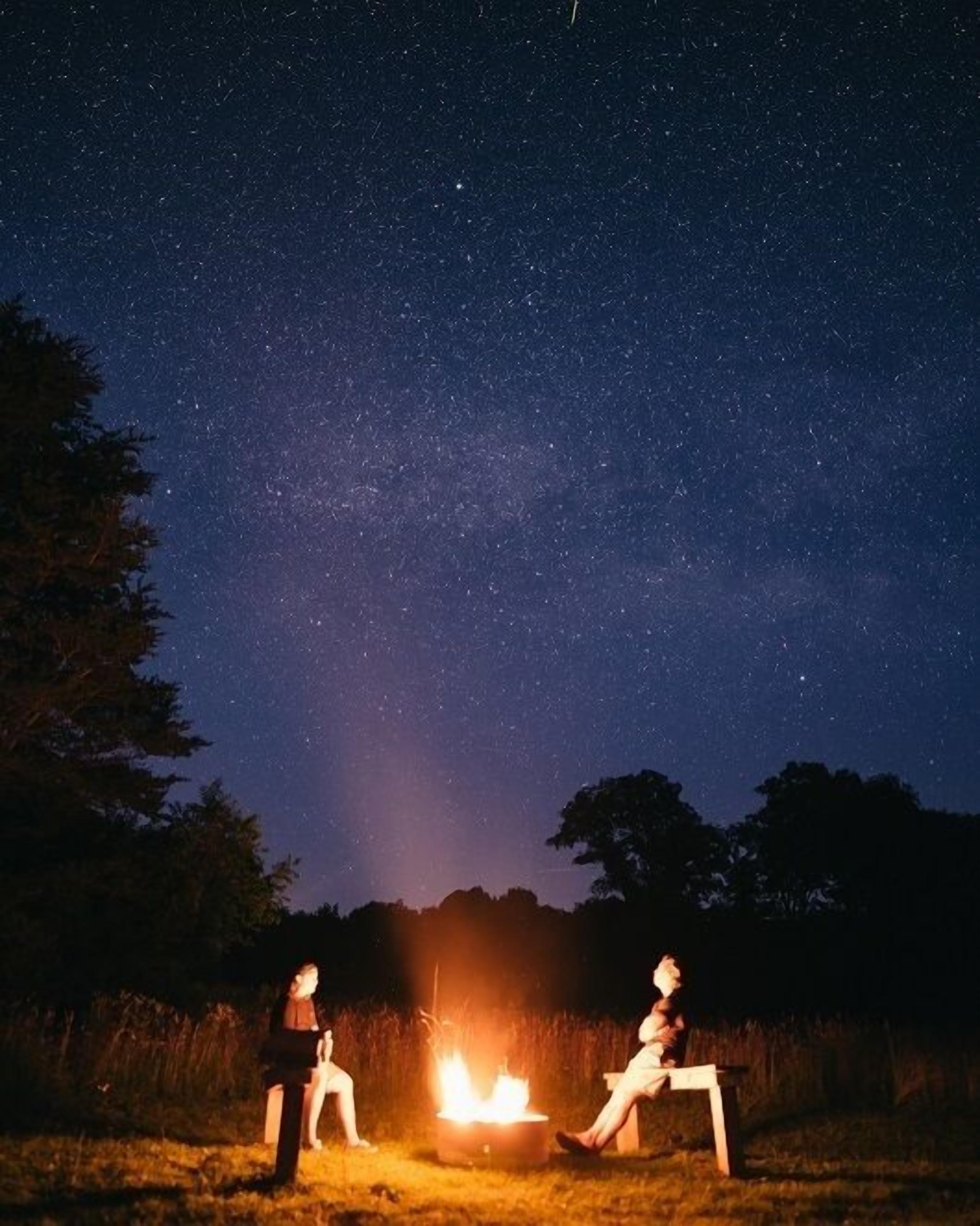 Dos personas sentadas junto a una fogata bajo un cielo estrellado.