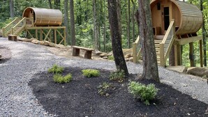 Kaksi puista leikkimökkiä, joissa on ulkoportaat, ja jotka sijaitsevat metsässä, jossa on sorapolku ja pieniä pensaita.