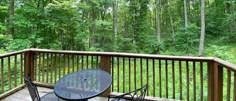 Terraza de madera con mesa y sillas de vidrio, con vistas a un bosque frondoso.