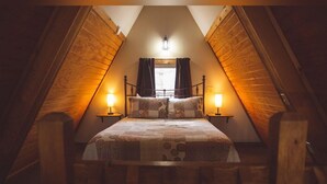 Een gezellig slaapkamer onder een schuin houten dak, een bed met een geborduurde quilt, twee bedlampjes en een raam met gordijnen.