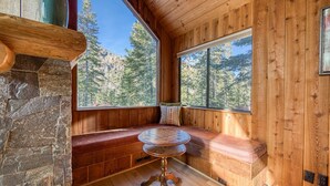 Interior de una cabaña de madera con chimenea de piedra, una mesa redonda de madera y un banco con vistas al bosque.