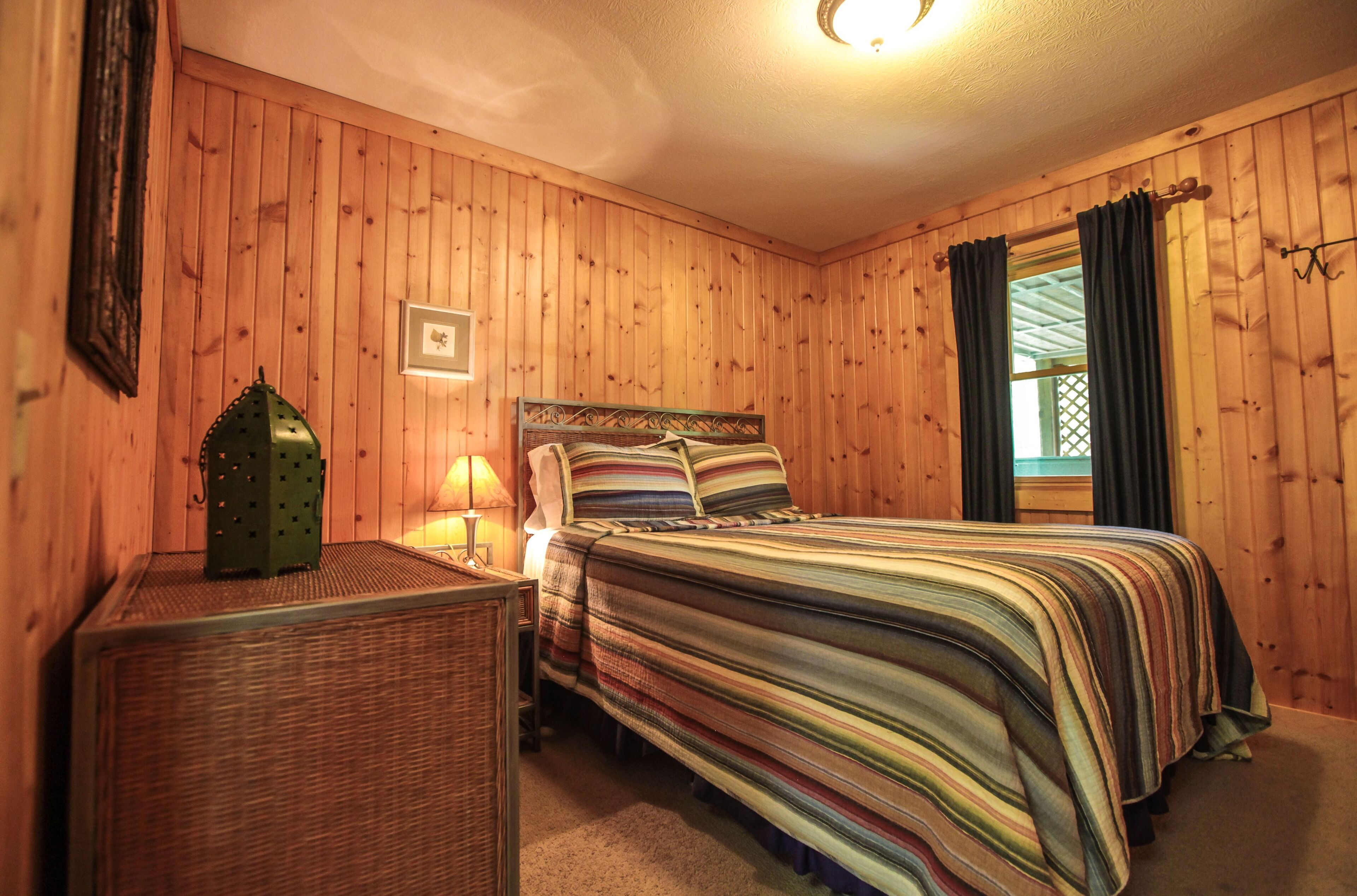 Ein Zimmer mit Holzwandverkleidung, einem Bett, einem Nachttisch, einer Lampe und einem Fenster mit Vorhängen.
