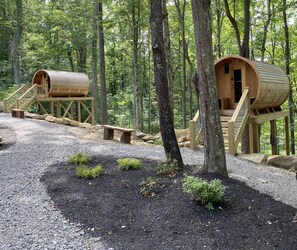 Deux cabanes en bois, reliées par des escaliers en bois, nichées dans une forêt où serpente un sentier de gravier, bordé de petits arbustes.
