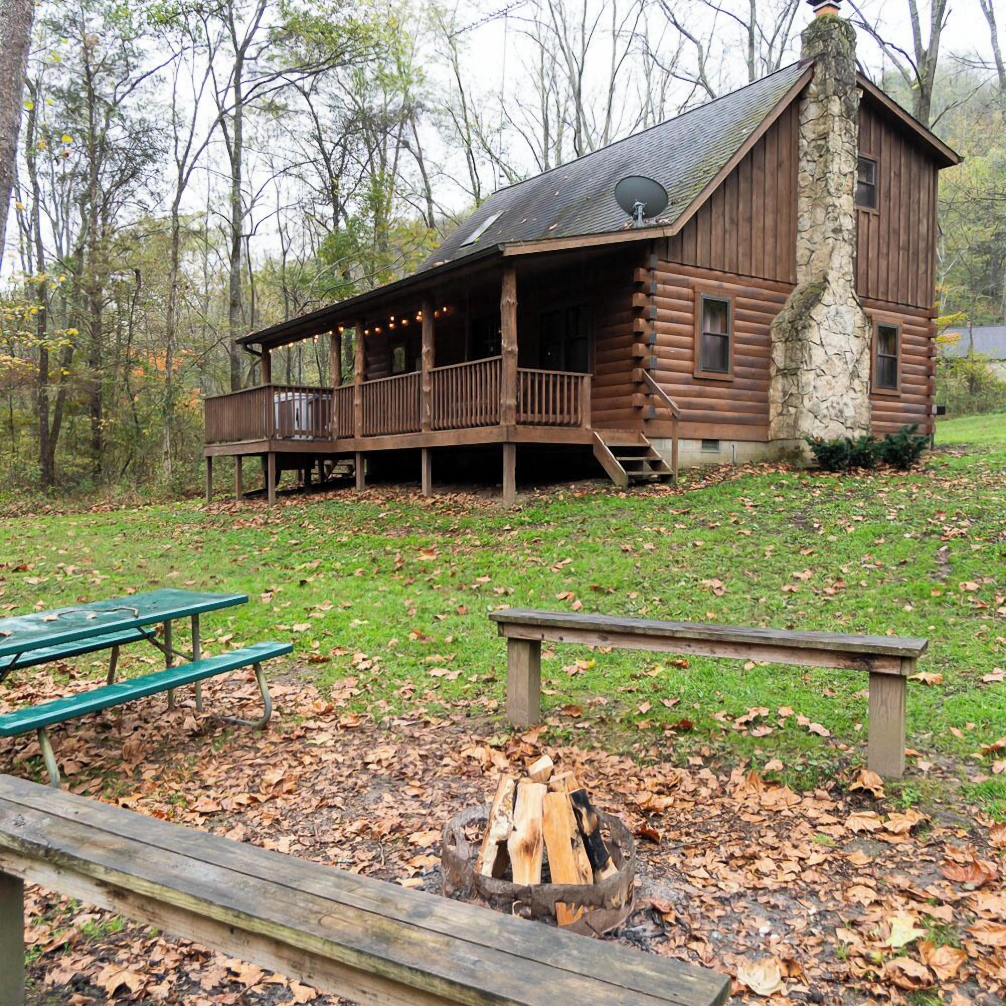A rustic log cabin with a porch and a fire pit in the foreground.