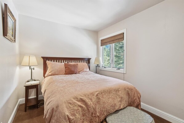 A bedroom with a bed, two bedside lamps, a window with a view of trees, and a picture on the wall.