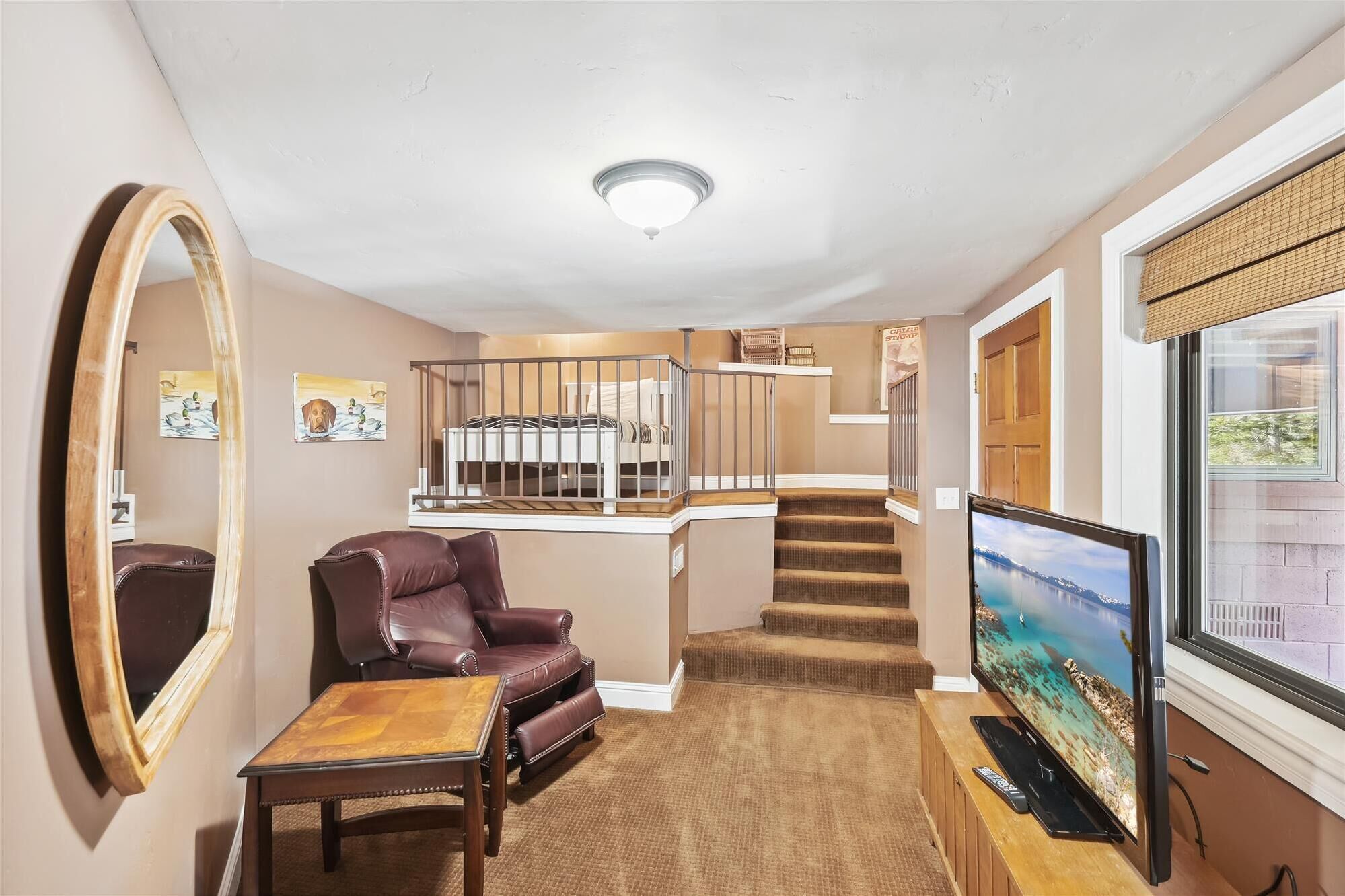 A living room with a brown leather sofa, a wooden coffee table, a large mirror, a television, and a staircase leading to a loft bed area.