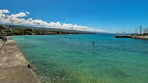 A coastal scene with clear turquoise water, a concrete walkway, and a distant mountain range under a blue sky with clouds. - Downtown Air Conditioned Condo: Walk to Beaches (Kailua-Kona)