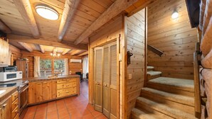Interior de una cabaña de madera con cocina, escaleras de madera y un ventanal grande.