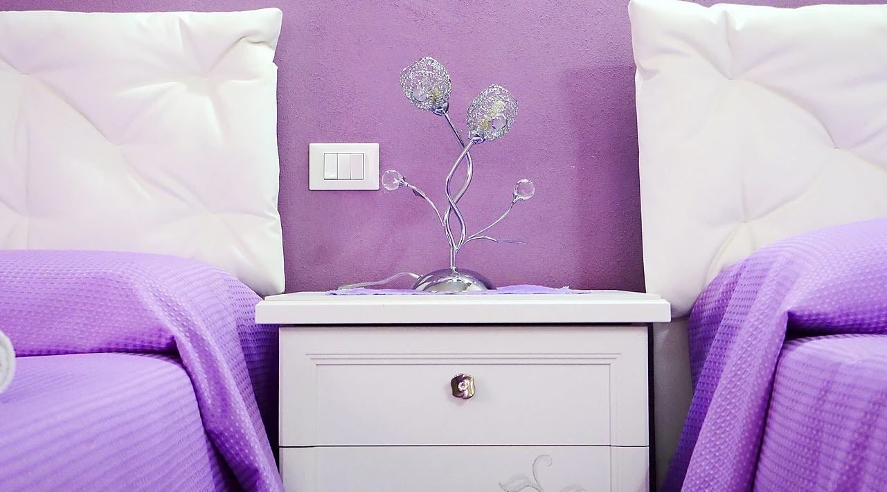 Un dormitorio con ropa de cama morada, una mesita de noche blanca con una flor de vidrio decorativa y una pared morada.