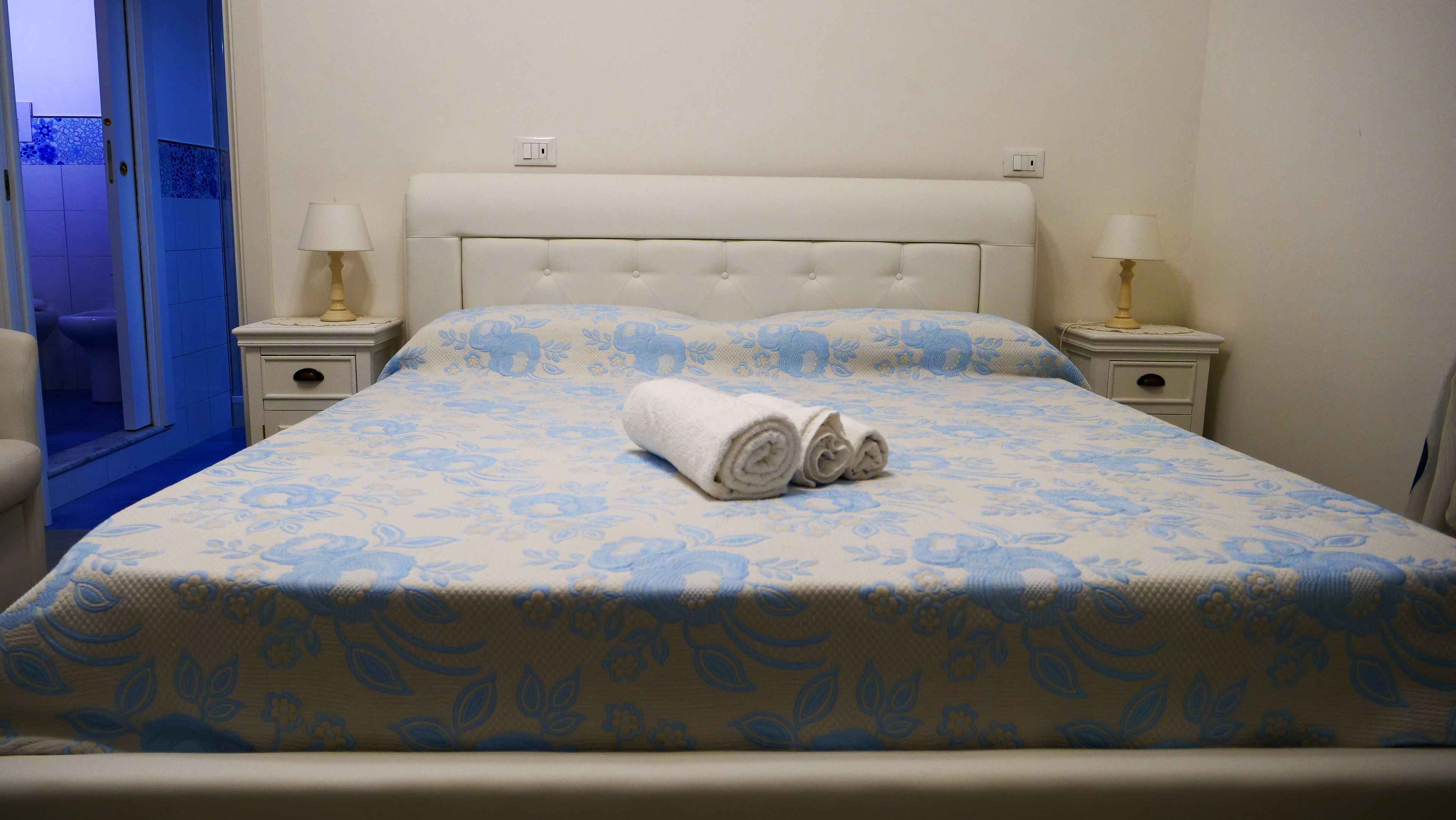Un letto rifatto con una coperta a fiori, due abat-jour e un asciugamano arrotolato sul letto.
