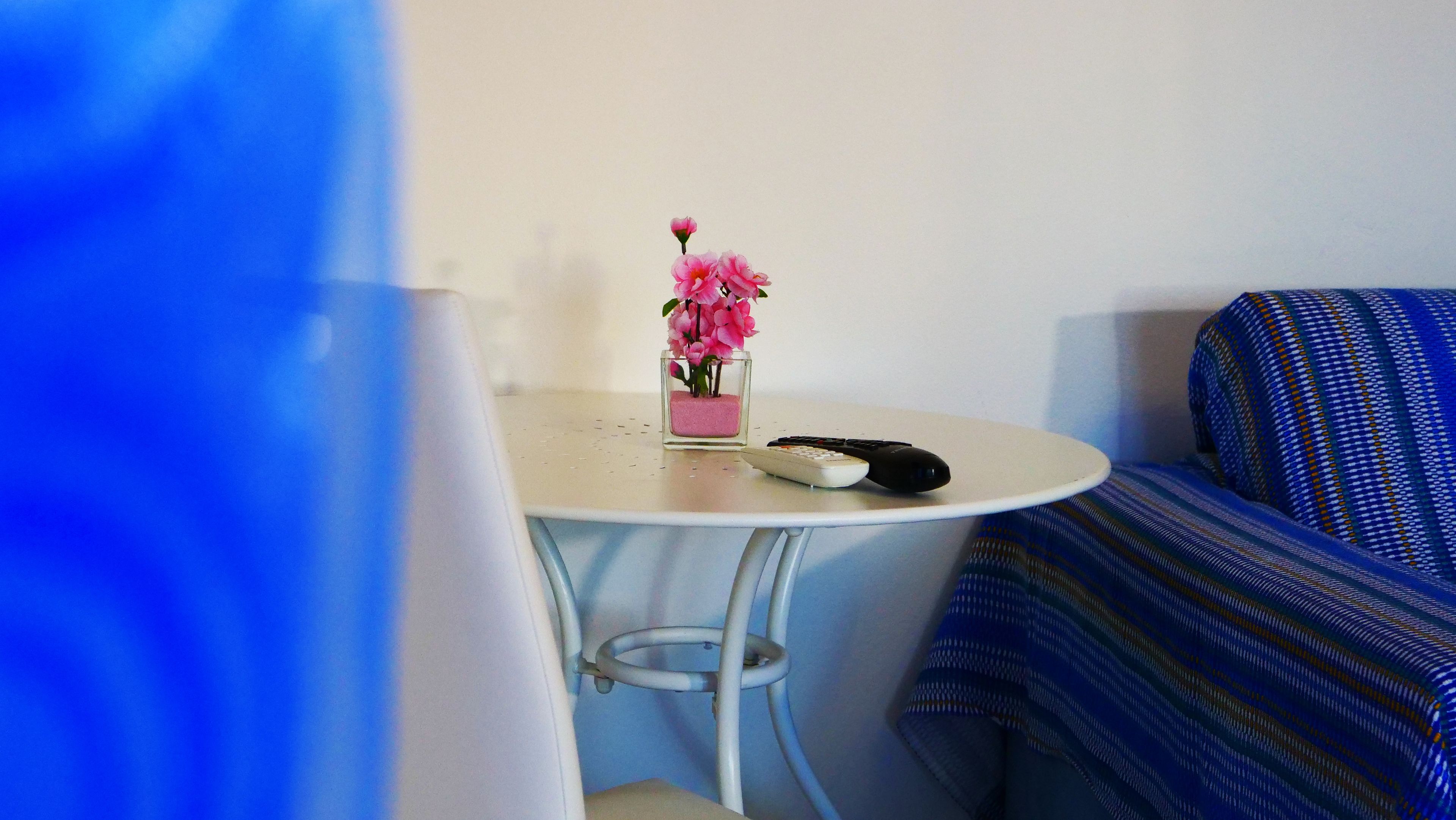 Une petite table ronde sur laquelle se trouve un vase rempli de fleurs roses, une télécommande et un lit recouvert d’une couverture à motifs bleus.