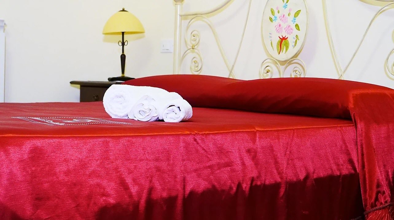 A neatly made bed with a red bedspread and three rolled white towels.