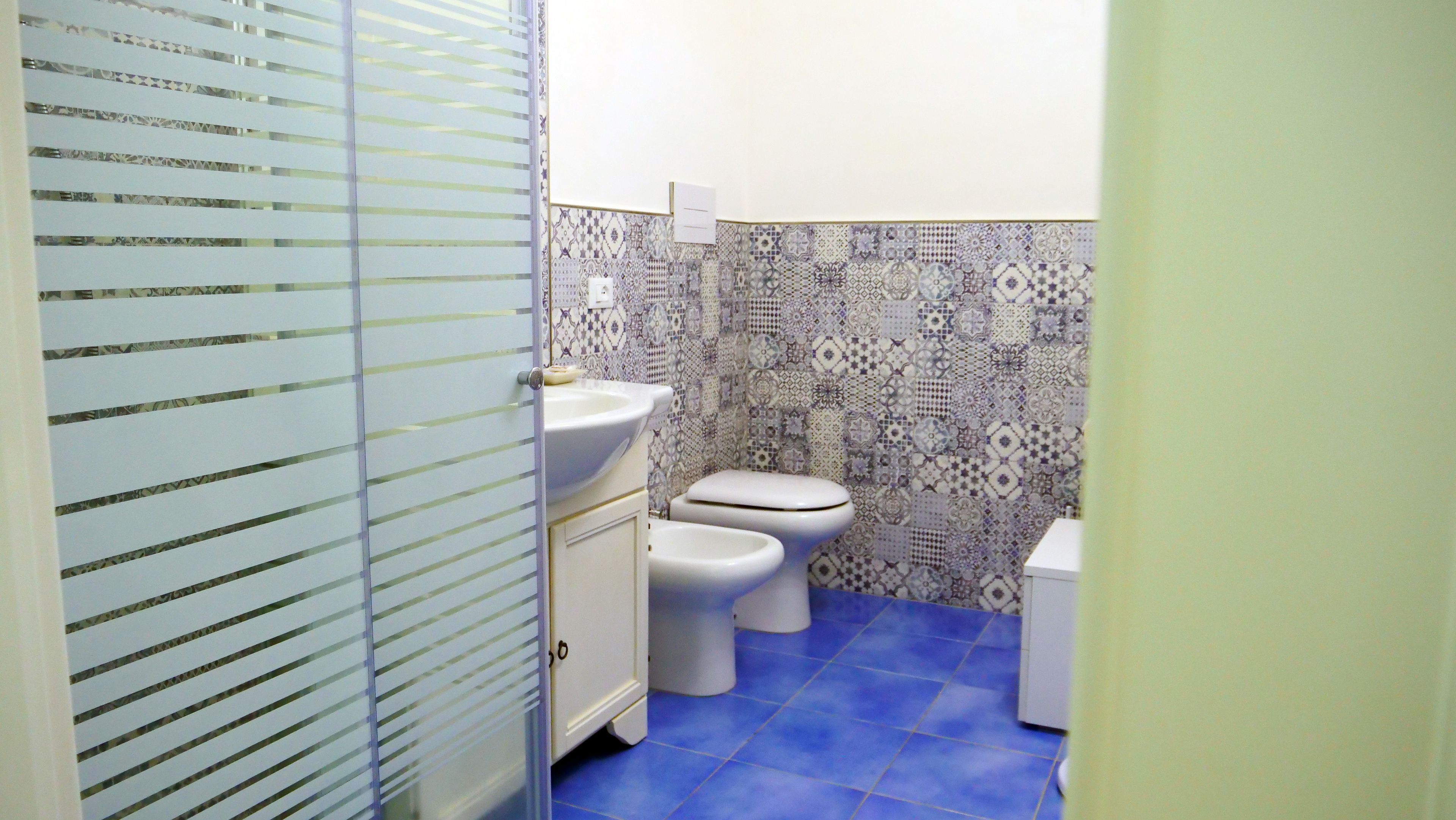 Ein Badezimmer mit Toilette, Bidet und Waschbecken. Der Boden ist blau gefliest, die Wände mit einem gemusterten Fliesenbelag versehen.