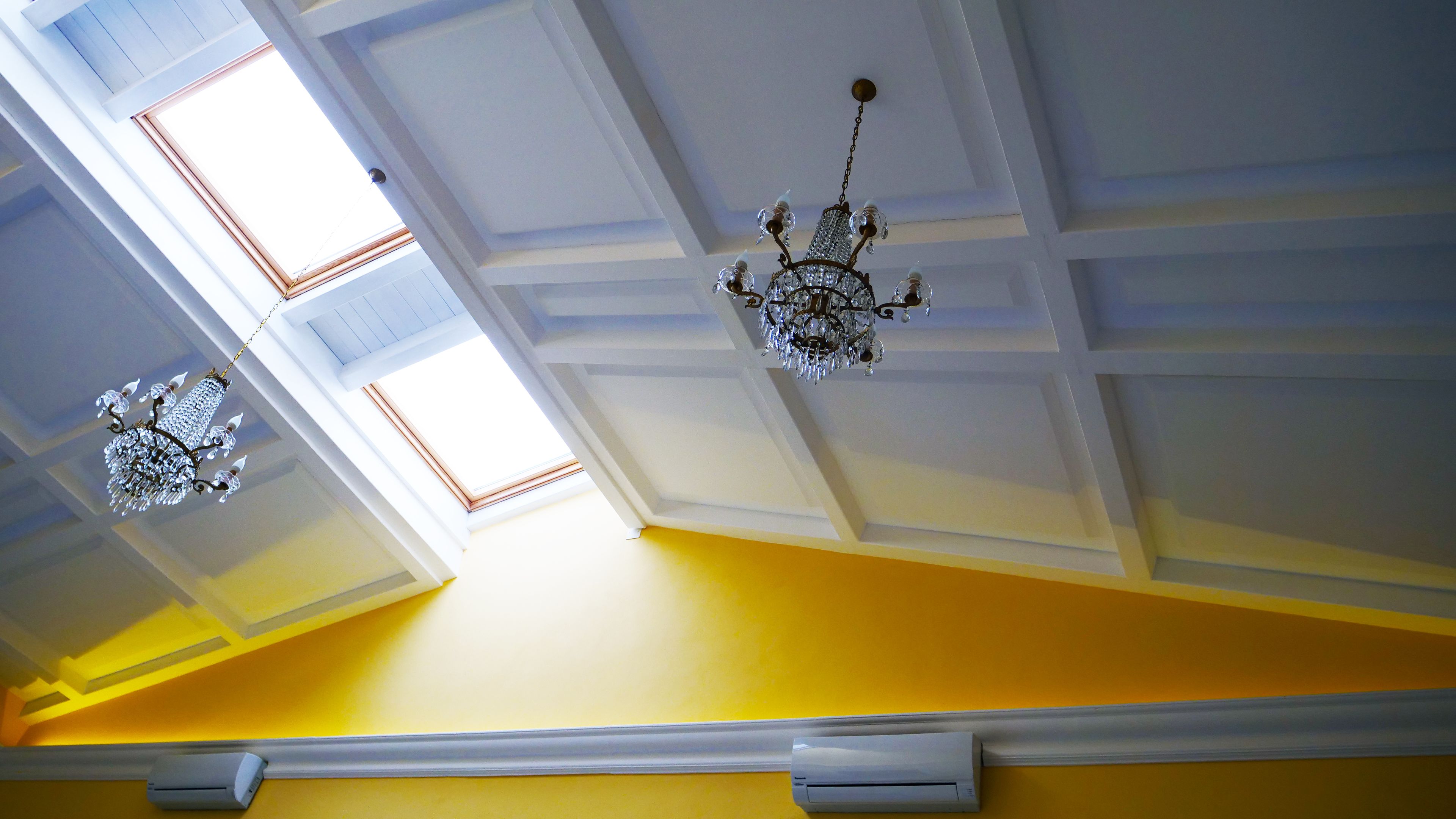 A room with a yellow wall, a white ceiling with skylights, and two chandeliers.