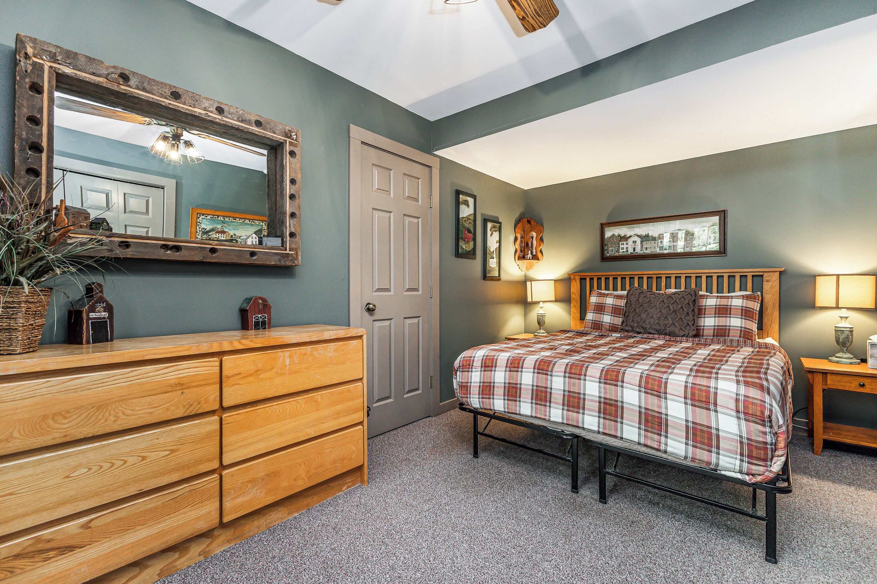 A bedroom with a wooden dresser, a bed with plaid bedding, a mirror, and a door.