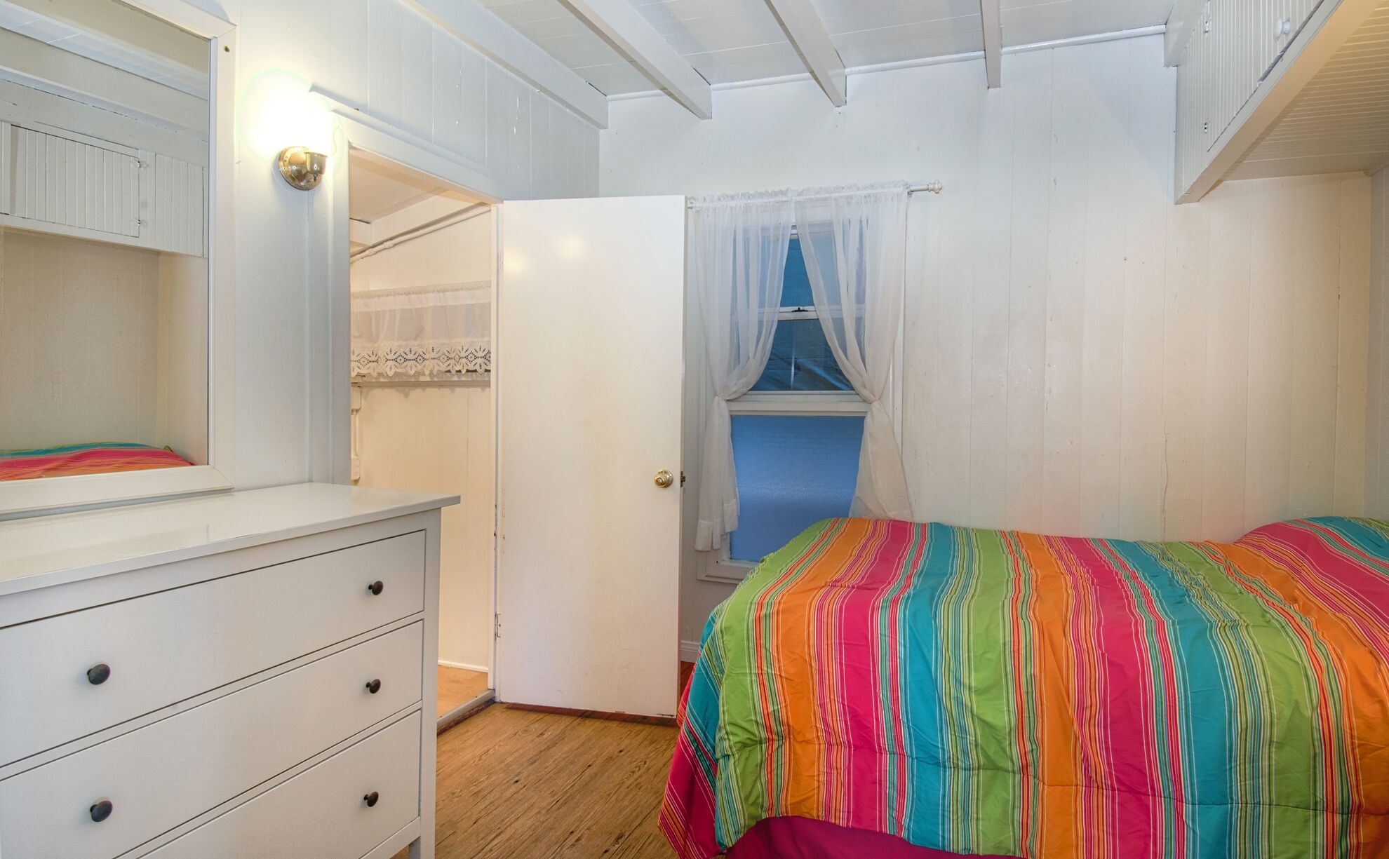 Een slaapkamer met een witte dressoir, een kleurrijk gestreept dekbedovertrek, een raam met dunne gordijnen en een houten vloer.
