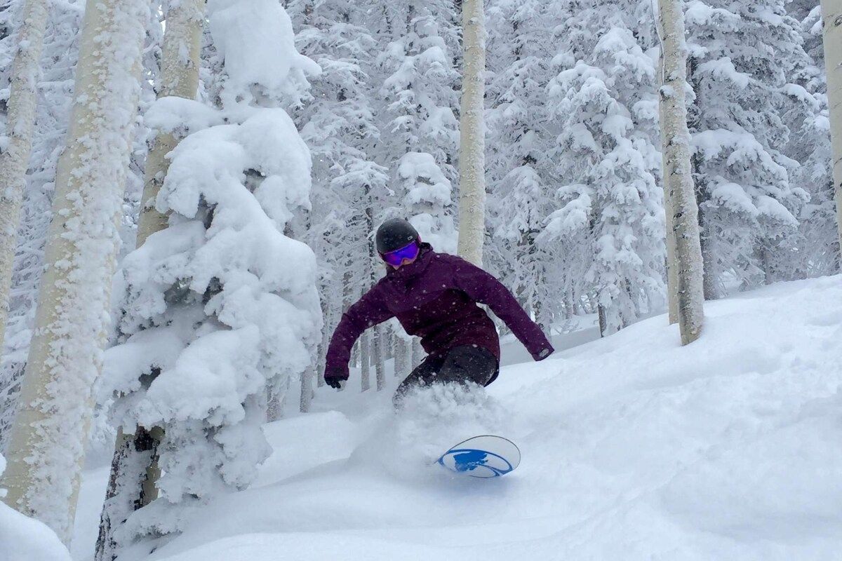 Una persona practica snowboard en una ladera nevada con árboles al fondo.