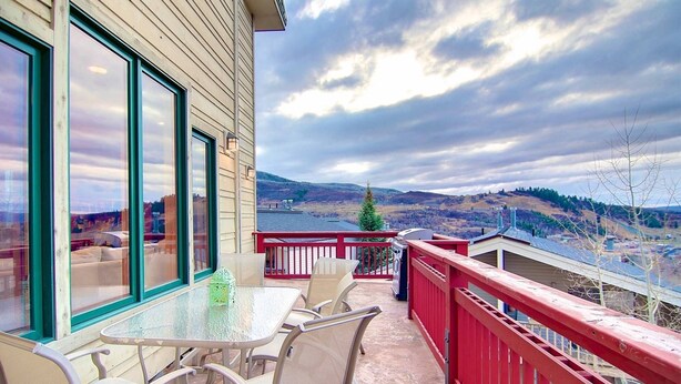 Un balcon avec une table et des chaises en verre, une balustrade rouge et une vue panoramique sur des collines et un lac.