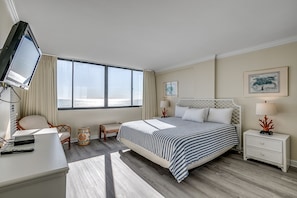Une chambre d’hôtel avec un grand lit, un bureau avec une télévision, une chaise, une table de chevet et une vue sur la mer.