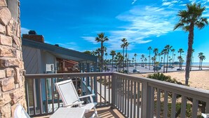 Ein Balkon mit weißen Stühlen und einer Steinsäule, der auf einen Strand mit Palmen und einem klaren, blauen Himmel blickt.