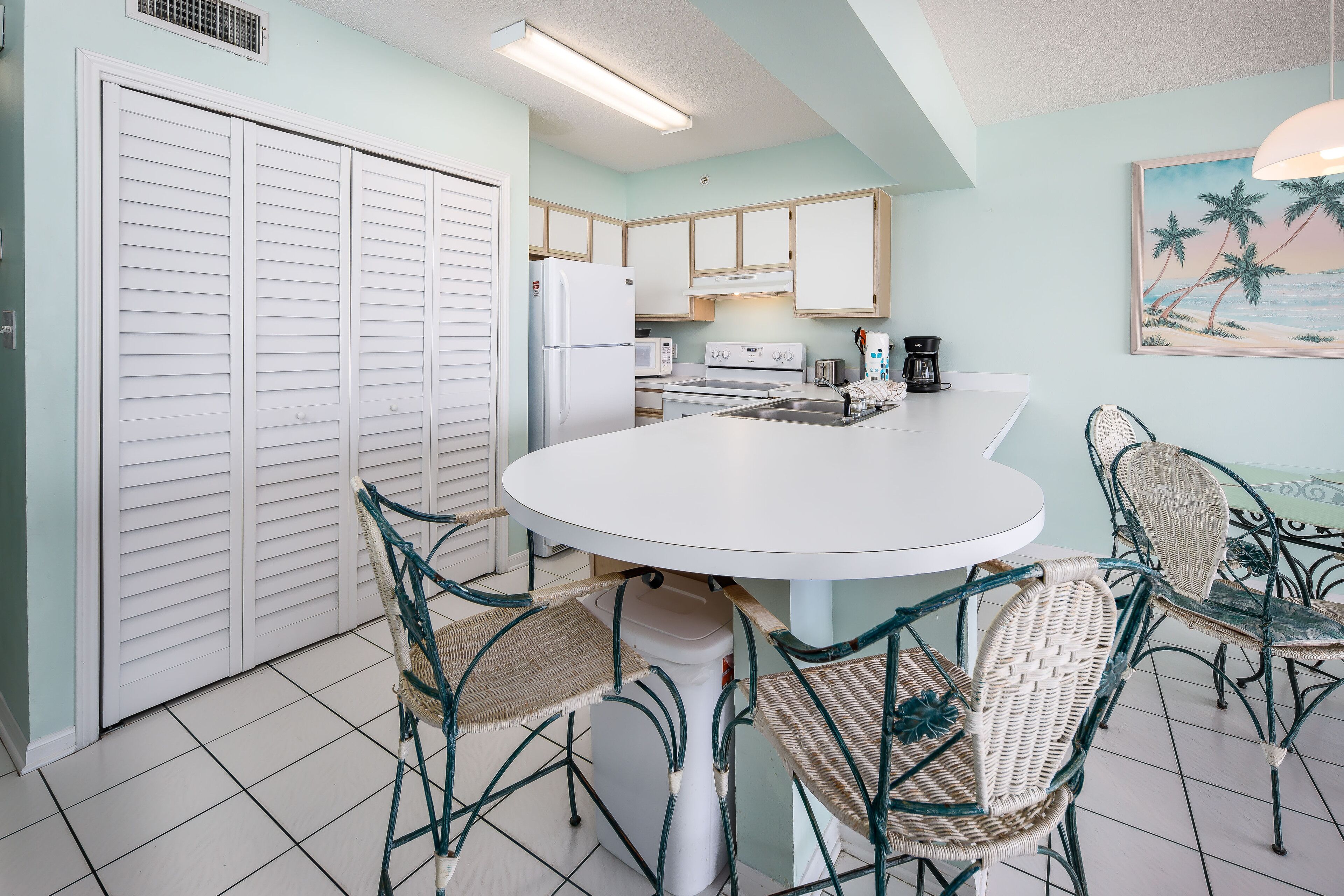 Une cuisine avec des meubles blancs, un réfrigérateur blanc, une cuisinière et un four à micro-ondes. On y trouve une table à manger avec des chaises en osier et un comptoir blanc.