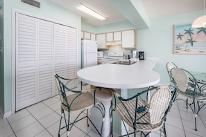 Une cuisine avec des meubles blancs, un réfrigérateur blanc, une cuisinière et un four à micro-ondes. On y trouve une table ronde entourée de quatre chaises en osier.