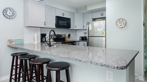 Une cuisine moderne avec des armoires blanches, un comptoir en granit et un réfrigérateur en acier inoxydable. On y trouve trois tabourets de bar et une horloge au mur.